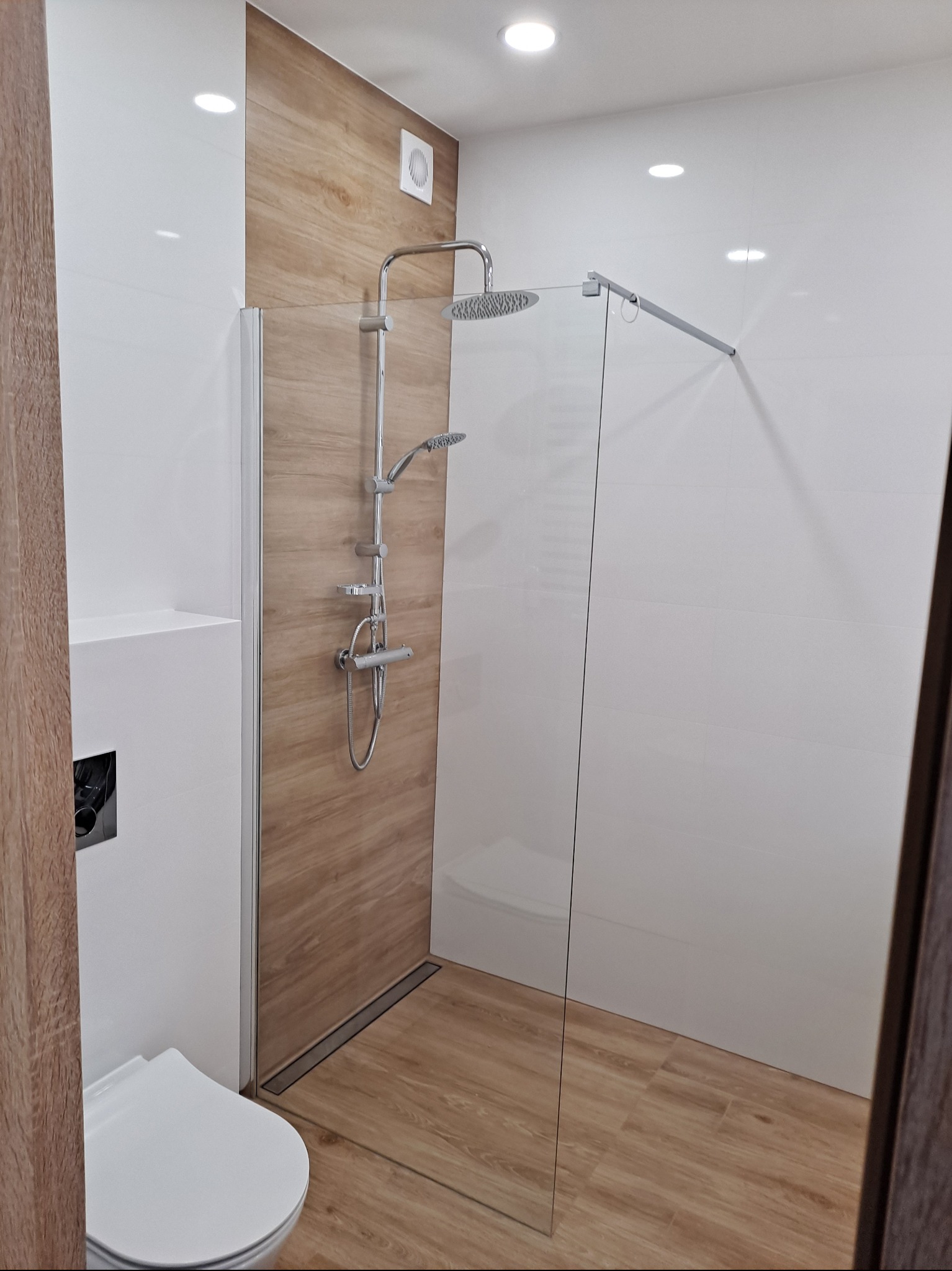 Nowoczesna łazienka z kabiną typu walk-in, wykończona białymi płytkami i panelami imitującymi drewno. Widoczny odpływ liniowy i minimalistyczna armatura.