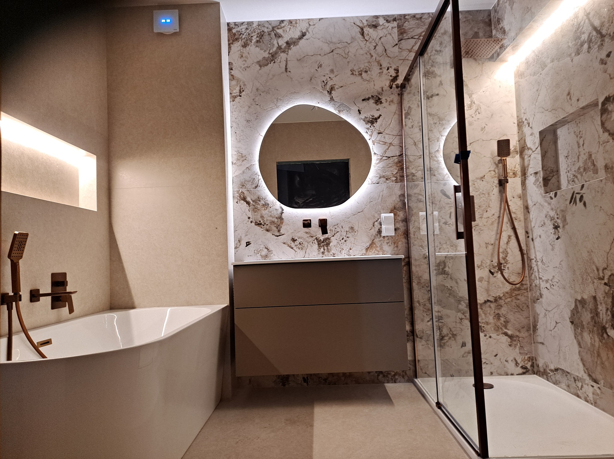 Nowoczesna łazienka w beżach: wanna, kabina prysznicowa, umywalka z podświetlanym lustrem o nieregularnym kształcie. Armatura w kolorze miedzi. Marmurowe płytki na ścianach.