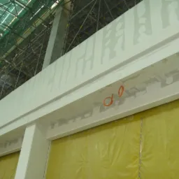 Sufit w trakcie prac wykończeniowych z widocznymi elementami konstrukcyjnymi, częściowo zakryty folią ochronną, zarysowane miejsca po łączeniach płyt. Widoczne rusztowania w tle.