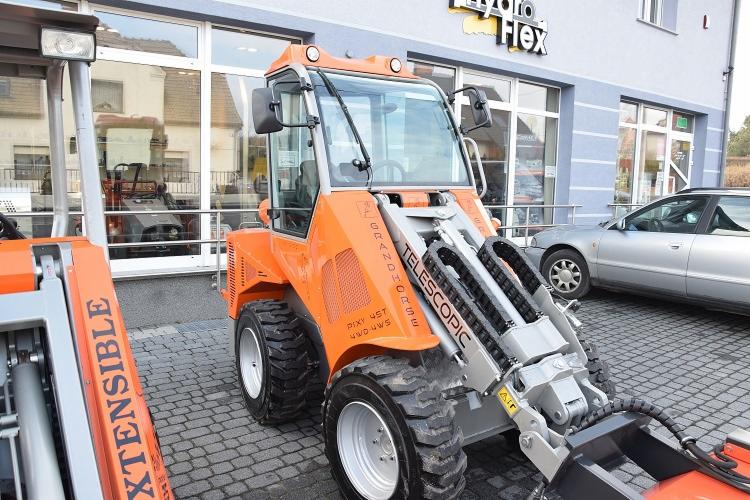 Pomarańczowa ładowarka teleskopowa Grandhorse Pixy 45T 4WD 4WS na tle budynku z dużymi oknami i zaparkowanym srebrnym samochodem.