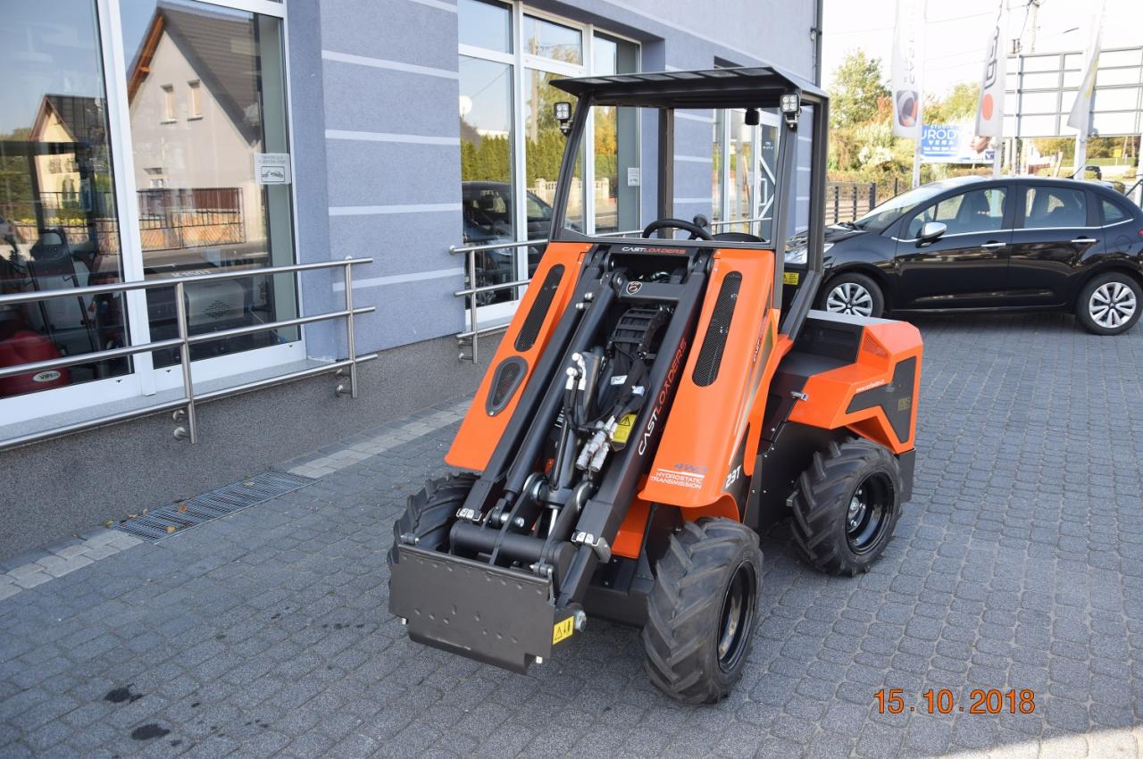 Pomarańczowa miniładowarka Castloaders 23T z czarną kabiną i oponami, zaparkowana na kostce brukowej przed budynkiem z dużymi oknami, w tle czarny samochód.