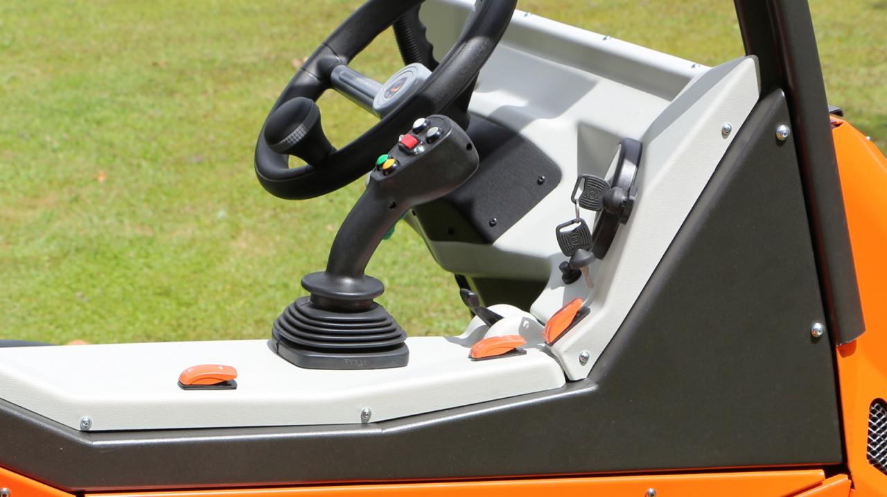 Wnętrze kabiny pomarańczowej maszyny budowlanej z joystickiem, kierownicą i przełącznikami na tle trawnika.