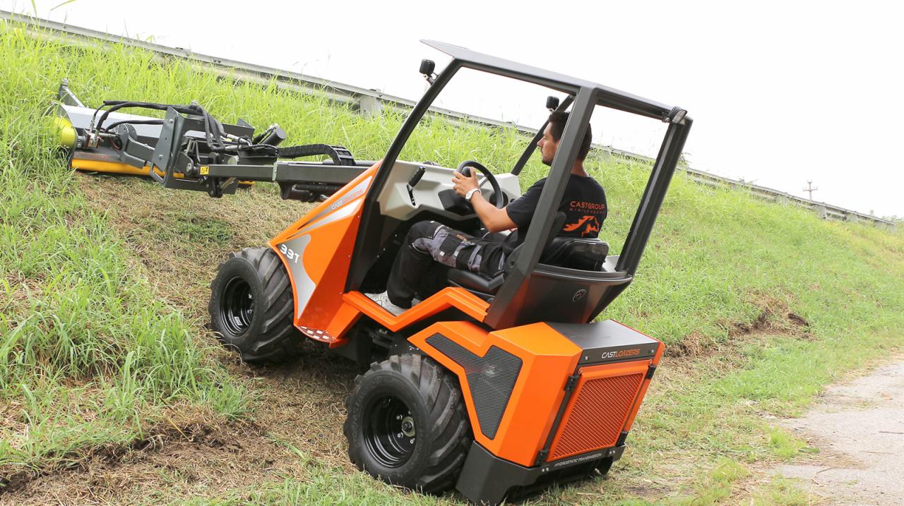 Pomarańczowa miniładowarka Castloaders 33T z operatorem podczas koszenia trawy na zboczu, widok z boku.