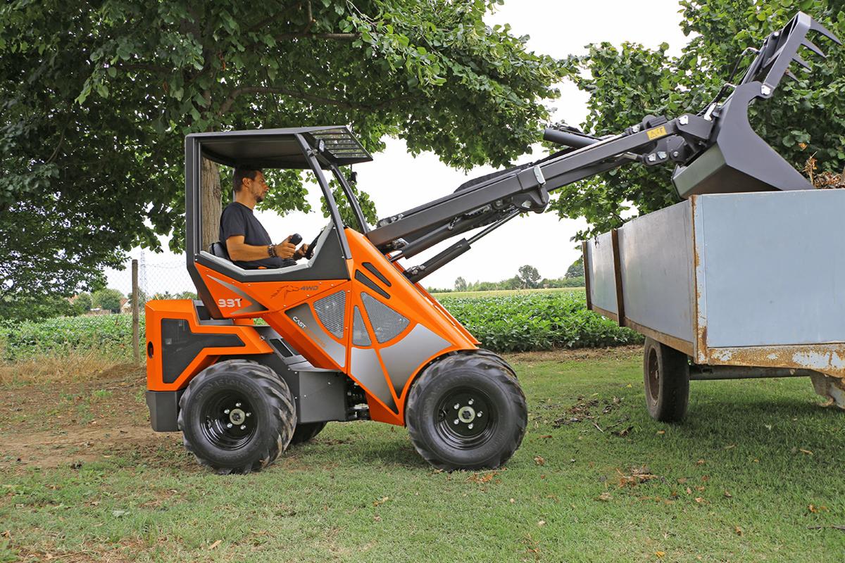 Pomarańczowa miniładowarka z operatorem, wysypująca gałęzie do przyczepy na tle zielonego pola i drzew.