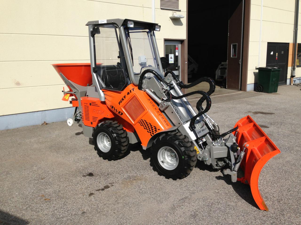 Pomarańczowa miniładowarka Pixy 41T z pługiem śnieżnym i zasobnikiem na posypkę, zaparkowana na zewnątrz budynku z otwartymi drzwiami garażowymi.