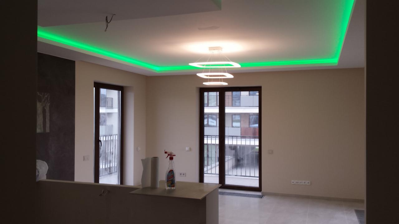 Nowoczesne wnętrze: podwieszany sufit z zielonym oświetleniem LED, minimalistyczna lampa, okna balkonowe. Widok na budynek z zewnątrz.