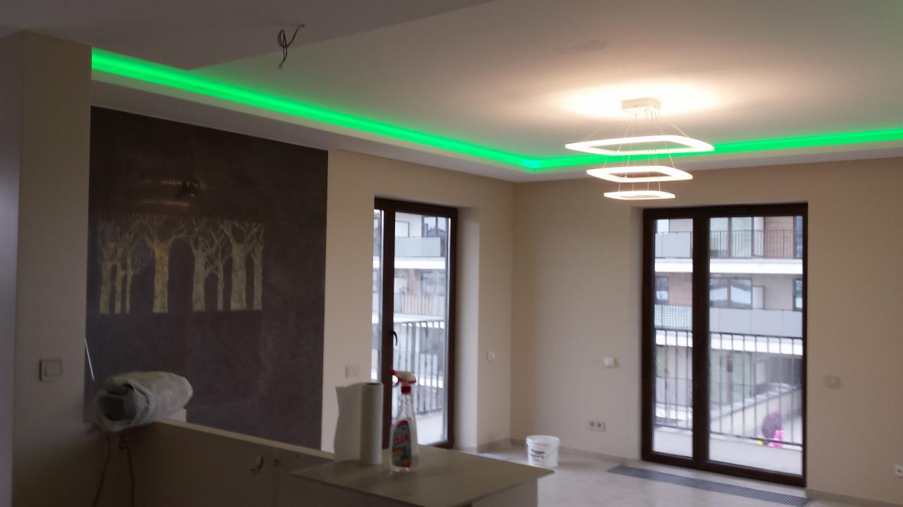 Nowoczesne wnętrze z zielonym oświetleniem LED podsufitowym, designerską lampą i ścianą z motywem drzew. Widok na balkon przez duże okna.