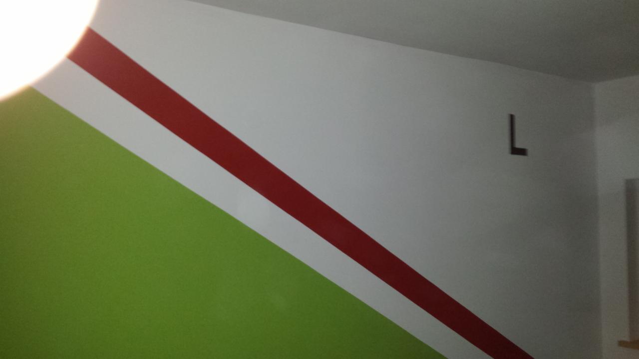 Ściana z ukośnymi pasami: zielony, biały i czerwony. Na białej części ściany wisi mała, ciemna litera L. Widok z dołu, z widocznym fragmentem sufitu.