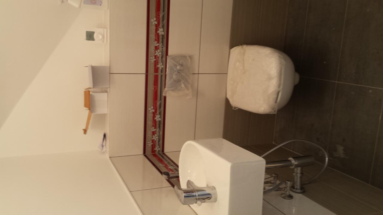 Nowa, biała umywalka z chromowaną baterią i częściowo zakryta folią toaleta w wykończonej łazience z brązowymi i białymi płytkami z dekoracyjnym pasem.