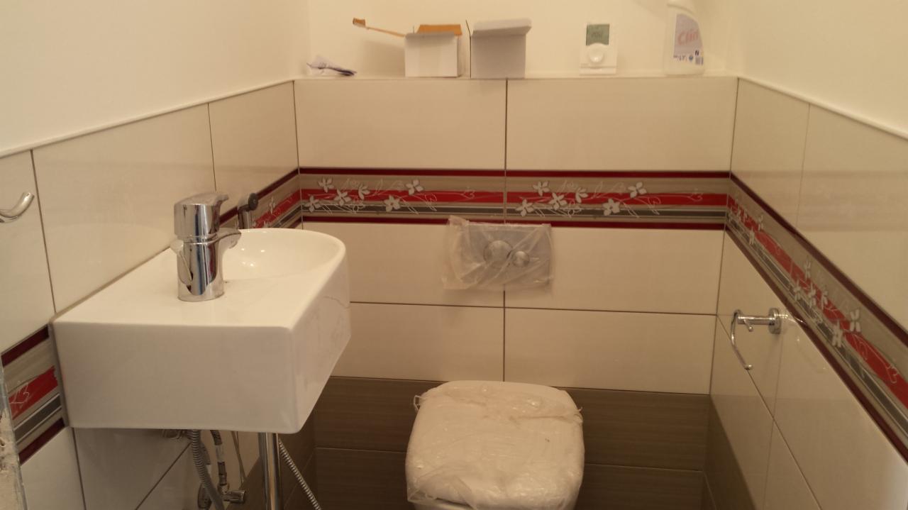 Wykończona łazienka z białą umywalką, baterią i toaletą w folii ochronnej. Kremowe płytki z dekoracyjnym pasem i brązowe płytki podłogowe. Nowoczesne wnętrze.