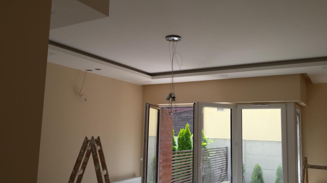 Wykończenie sufitu podwieszanego i ścian w beżowym odcieniu w trakcie prac remontowych w Warszawie. Widoczna drabina i niedokończone oświetlenie.