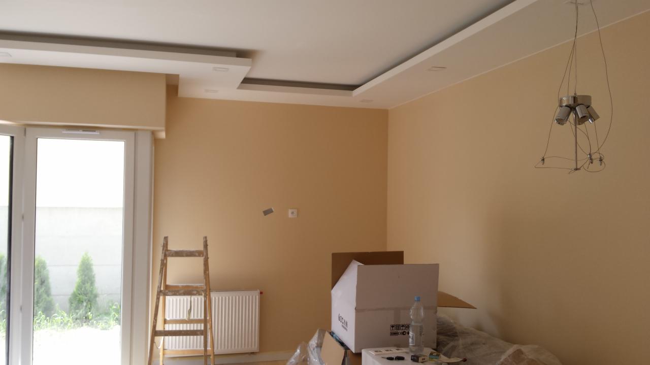 Beżowe ściany w pokoju po gładziowaniu, widoczna drabina, grzejnik, kartony i lampa wisząca. Wykończenie sufitu z oświetleniem punktowym.