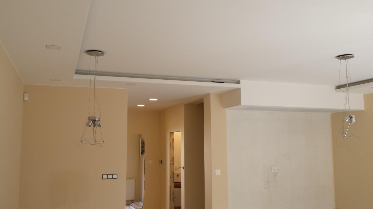 Jasne wnętrze po remoncie: gładkie ściany w beżowym odcieniu, podwieszany sufit z oświetleniem punktowym i wiszącymi lampami. Widok na sąsiednie pomieszczenie.