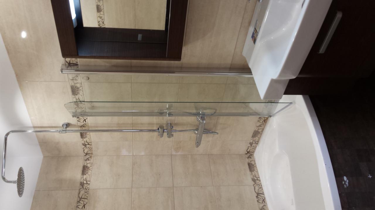 Wykończona łazienka z beżowymi płytkami, kabiną prysznicową ze szklanym panelem i deszczownicą. Biała umywalka z szafką i lustro w ciemnej ramie.