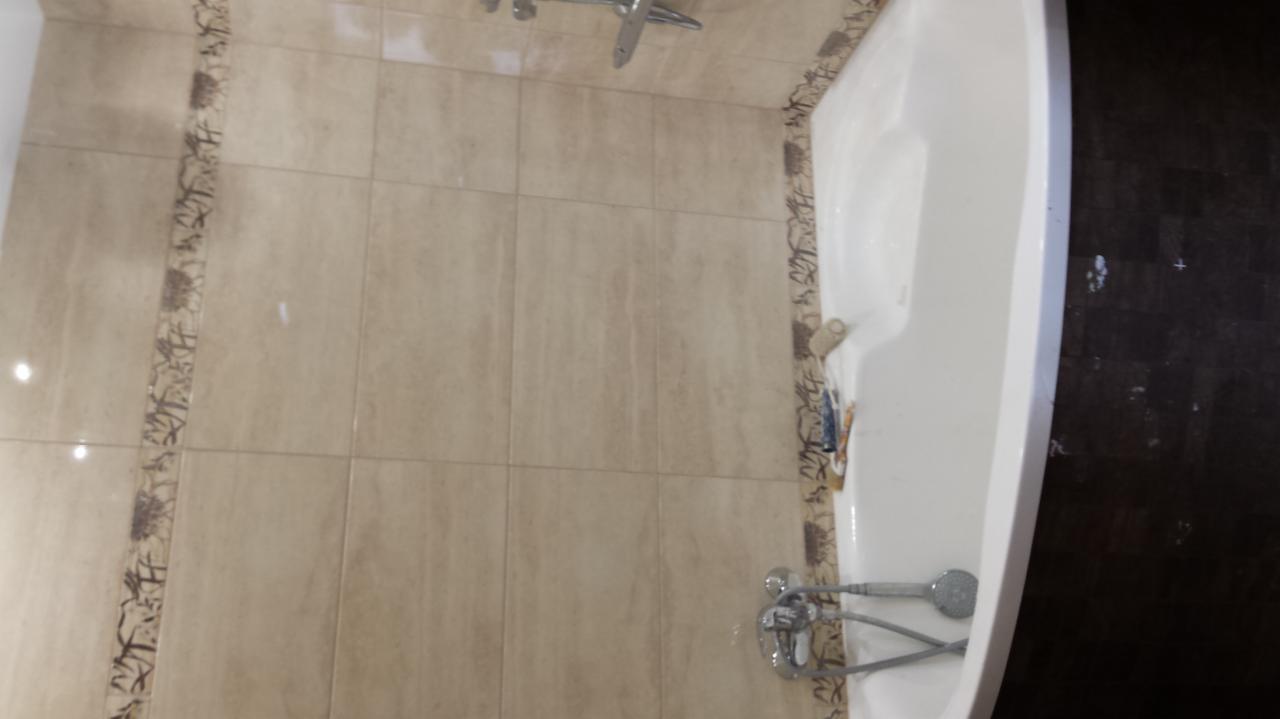 Wykończona łazienka z beżowymi płytkami i dekoracyjnym pasem. Biała wanna z chromowaną armaturą. Widok na fragment ściany i wanny.