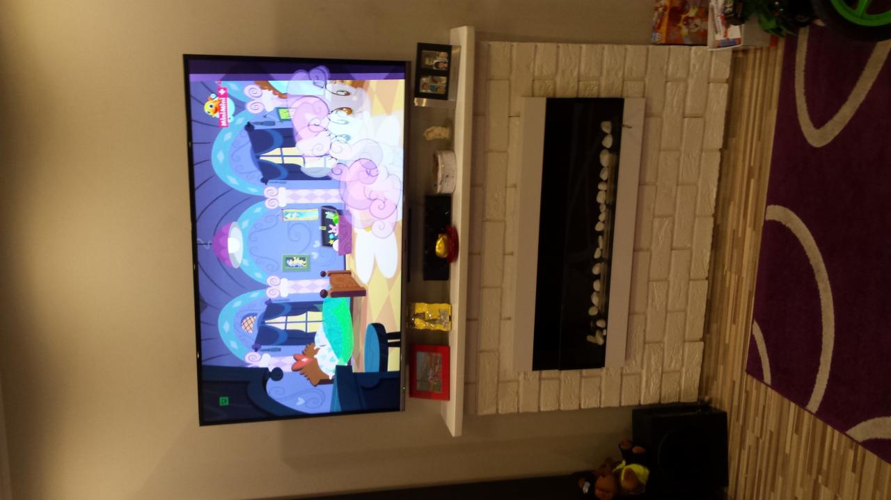 Biały, ceglany kominek z czarną wnęką i białymi kamieniami dekoracyjnymi, obok telewizor z włączoną bajką, na ścianie półka z dekoracjami, dywan w tle.