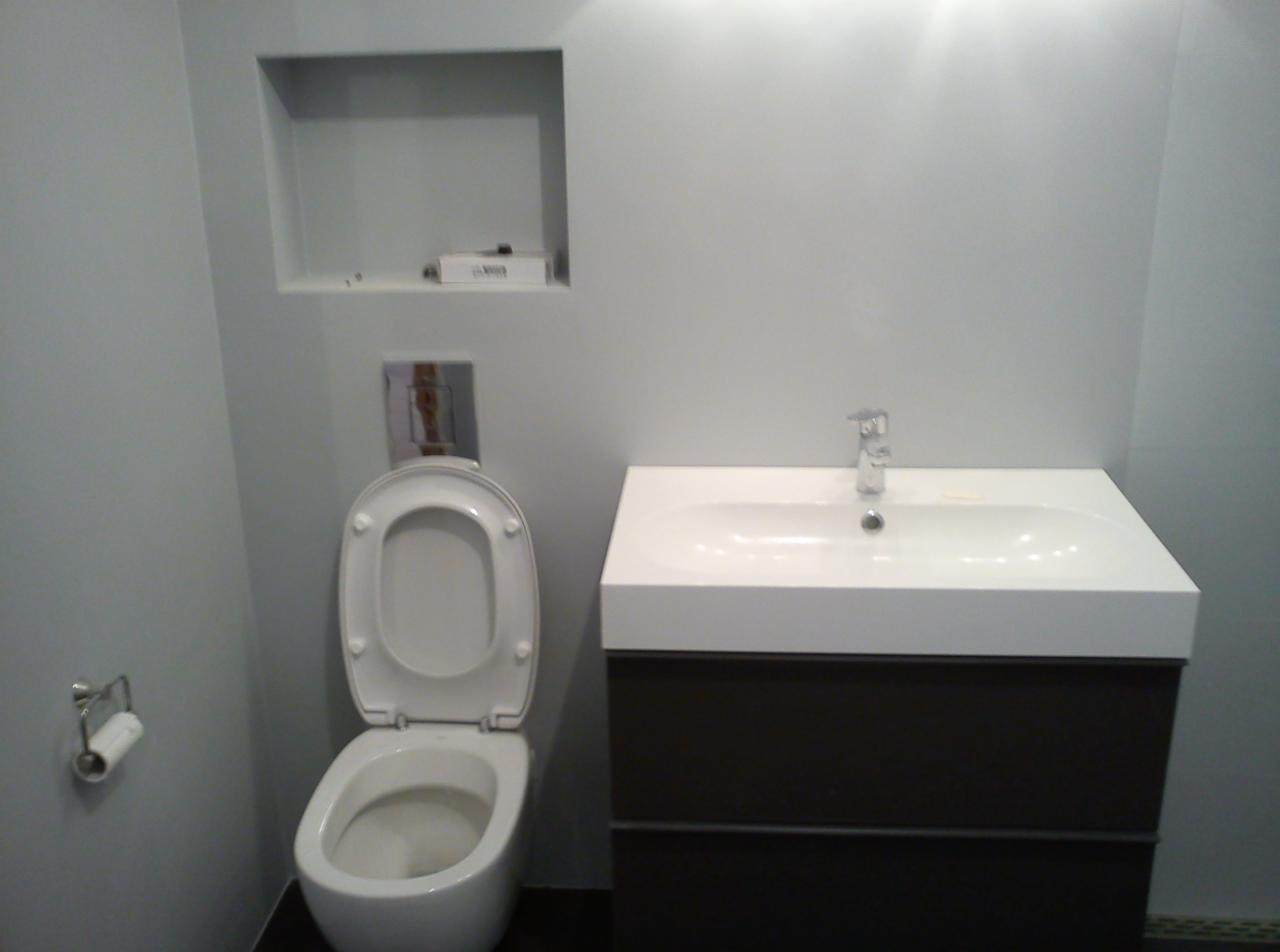 Wykończona łazienka w szarościach: toaleta z otwartą klapą, biała umywalka na szafce w kolorze ciemnego brązu, wnęka w ścianie. Minimalistyczny design.