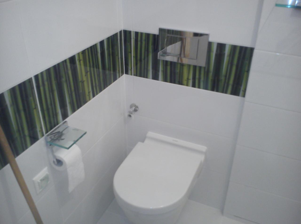 Biała toaleta podwieszana w łazience z akcentem bambusowych płytek dekoracyjnych. Szklana półka nad rolką papieru toaletowego. Nowoczesny design, czystość i minimalizm.