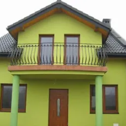 Dom z zieloną elewacją i balkonem z ozdobną, czarną balustradą. Widoczne brązowe okna i drzwi. Dach pokryty szarą dachówką.