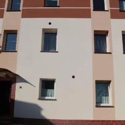 Wielobarwna elewacja budynku wielorodzinnego z widocznymi oknami i wejściem z daszkiem. Zastosowano różne odcienie beżu i brązu, tworząc geometryczny wzór.