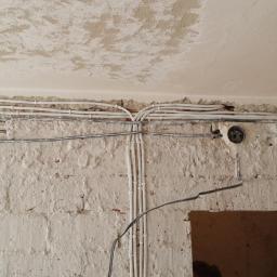 Przemysław Stawarski - Surowa ściana z widocznymi przewodami elektrycznymi w białych rurkach, prowadzącymi wzdłuż ściany i pod sufitem, z fragmentami starego tynku na suficie i otworem w murze w prawym dolnym rogu.