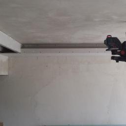 Przemysław Stawarski - Wnętrze w trakcie remontu z widoczną konstrukcją sufitu podwieszanego, poziomica laserowa na statywie oraz stara lampa wisząca.