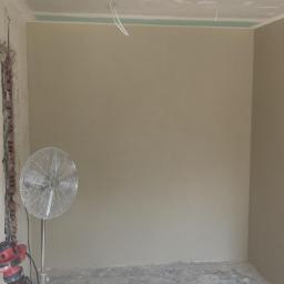 Przemysław Stawarski - Pokój po nałożeniu gładzi gipsowej na ściany i sufity, z widoczną instalacją elektryczną, wentylatorem stojącym i bruzdą w ścianie po prawej stronie.