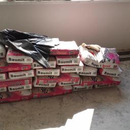 Przemysław Stawarski - Stos worków z zaprawą budowlaną Baumit Rapido 1 Speed na podłodze w surowym pomieszczeniu przed rozpoczęciem prac wykończeniowych, widoczne przewody instalacji elektrycznej w ścianie.