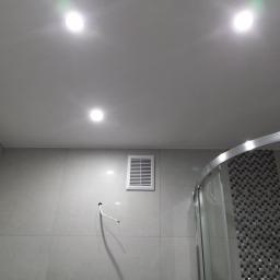 Przemysław Stawarski - Wykończona łazienka z szarą, kwadratową płytką na ścianach, sufit podwieszany z trzema punktami świetlnymi, wentylator w ścianie i fragment kabiny prysznicowej z mozaiką wewnątrz.