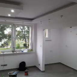 Wnętrze pokoju po remoncie z świeżo pomalowanymi ścianami na biało, szarą podłogą i oknem z widokiem na zieleń, widoczne narzędzia i akcesoria malarskie w rogu pomieszczenia.