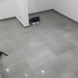 Przemysław Stawarski - Wykończona podłoga z szarych, kwadratowych płytek w jasnym pomieszczeniu, widoczny fragment worka z klejem i plecak w rogu pokoju.