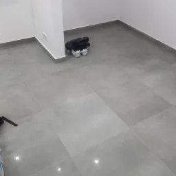 Wykończona podłoga z szarych, kwadratowych płytek w jasnym pomieszczeniu, widoczny fragment worka z klejem i plecak w rogu pokoju.