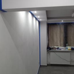 Przemysław Stawarski - Świeżo pomalowane na biało ściany z niebieską taśmą malarską zabezpieczającą sufit, widoczne halogeny w suficie, okno zasłonięte folią malarską, grzejnik i parapet z akcesoriami malarskimi.