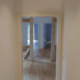 Korytarz po remoncie z jasną drewnianą podłogą, białe ściany i ościeżnice, widoczne drewniane podpory w przejściu do kolejnych pomieszczeń.