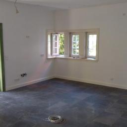 Przemysław Stawarski - Wnętrze pokoju po remoncie, z białymi ścianami, ciemną kamienną podłogą, oknem narożnym z otwartym skrzydłem, zielonymi framugami drzwi i widocznymi instalacjami elektrycznymi na ścianie.