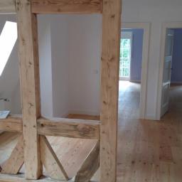 Przemysław Stawarski - Wnętrze domu w trakcie remontu, widoczne drewniane elementy konstrukcyjne, świeżo położona drewniana podłoga, białe ściany i otwarte drzwi do kolejnych pomieszczeń.