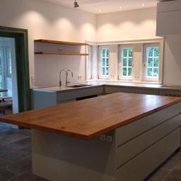 Przemysław Stawarski - Nowoczesna kuchnia z drewnianym blatem wyspy, szarozielonymi szafkami i kamienną podłogą, widok na jadalnię przez zielone drzwi.