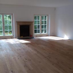 Przemysław Stawarski - Jasne, przestronne wnętrze z drewnianą podłogą, białymi ścianami i trzema oknami z widokiem na zieleń. Centralnym punktem jest kominek z kamienną obudową. Podłoga błyszczy w promieniach słońca.