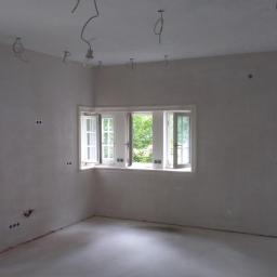 Przemysław Stawarski - Pokój w trakcie przygotowań do malowania: ściany po szpachlowaniu, widoczne puszki elektryczne, wiszące kable oświetleniowe, otwarte okno z widokiem na zieleń, drzwi z zielonymi futrynami.