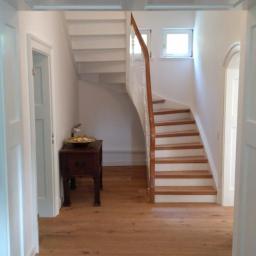 Przemysław Stawarski - Jasne wnętrze domu z drewnianą podłogą, schodami z drewnianymi stopniami i białymi balustradami, widoczne drzwi i mały stolik z dekoracją.