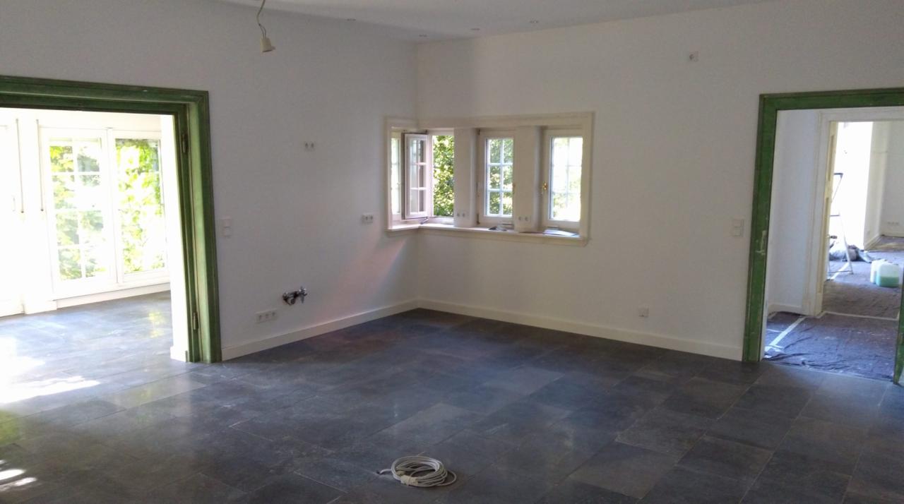 Wnętrze pokoju po remoncie, z białymi ścianami, ciemną kamienną podłogą, oknem narożnym z otwartym skrzydłem, zielonymi framugami drzwi i widocznymi instalacjami elektrycznymi na ścianie.