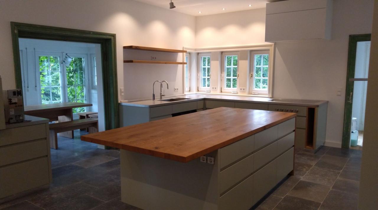 Nowoczesna kuchnia z drewnianym blatem wyspy, szarozielonymi szafkami i kamienną podłogą, widok na jadalnię przez zielone drzwi.