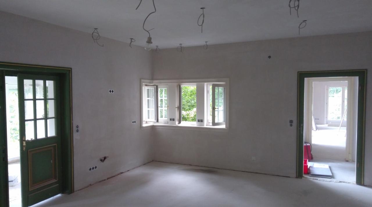 Pokój w trakcie przygotowań do malowania: ściany po szpachlowaniu, widoczne puszki elektryczne, wiszące kable oświetleniowe, otwarte okno z widokiem na zieleń, drzwi z zielonymi futrynami.