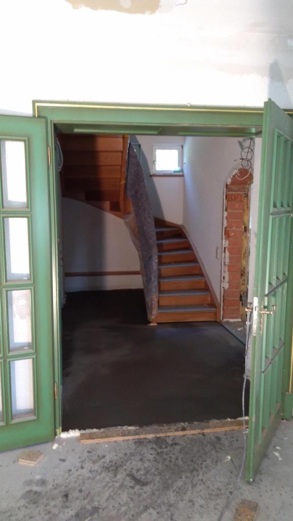 Wnętrze w trakcie remontu: świeżo wylana wylewka podłogowa, drewniane schody zabezpieczone folią, widoczne otwarte, zielone drzwi z szybkami i fragment ściany z odsłoniętą czerwoną cegłą.