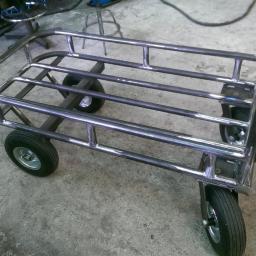Salda-col - Wózek transportowy wykonany ze spawanych rur stalowych, z czterema kołami, stojący na betonowej posadzce w warsztacie.