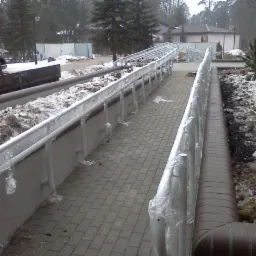 Zewnętrzna rampa dla wózków inwalidzkich z szarych kostek brukowych, zabezpieczona folią ochronną na metalowych poręczach, otoczona resztkami śniegu i ziemią.