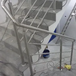 Metalowa balustrada schodowa z rur o szlifowanej powierzchni, zamontowana na betonowych schodach w trakcie prac wykończeniowych; widoczne przewody, drabina i wiadro z farbą.