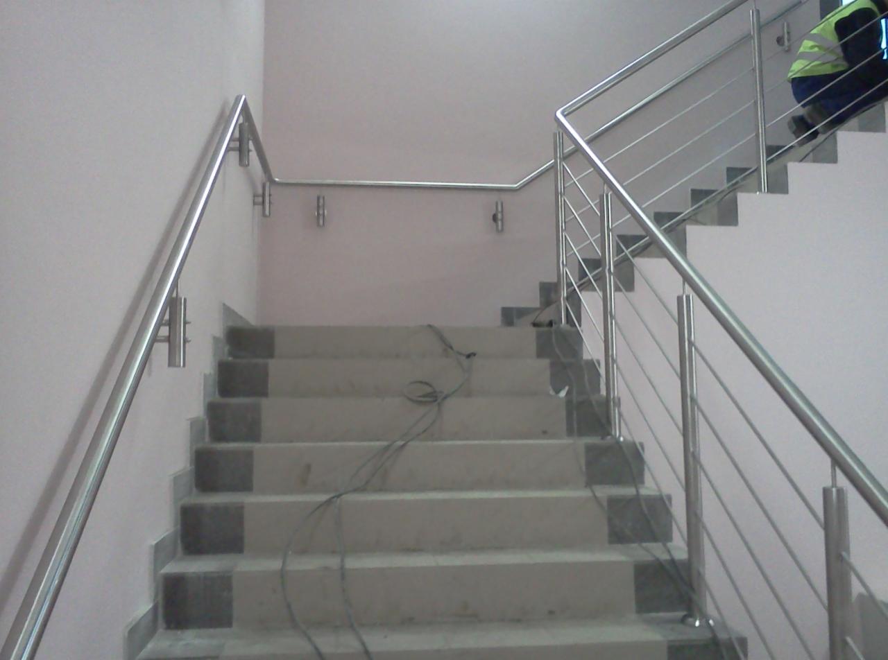 Stalowa balustrada schodowa z cienkimi prętami, montowana na jasnych schodach wewnątrz budynku, widoczny fragment osoby w odblaskowej kamizelce na górnych stopniach.