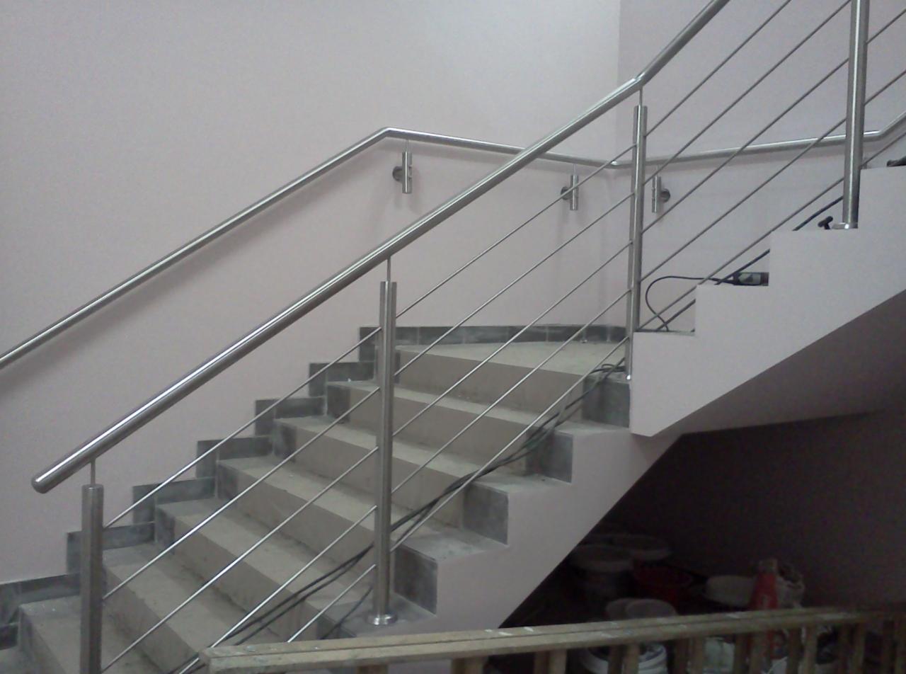 Klatka schodowa z nowoczesną, metalową balustradą ze stali nierdzewnej, zamontowaną na betonowych stopniach. Widoczne elementy mocujące do ściany i podłogi.