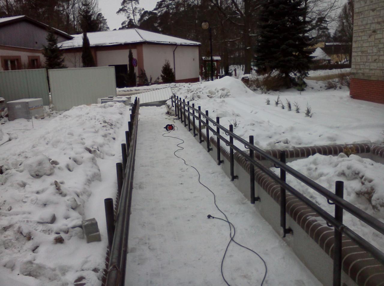 Zimowy montaż metalowej balustrady wzdłuż chodnika, widoczny przewód elektryczny na śniegu, w tle budynki i zaspy śnieżne.